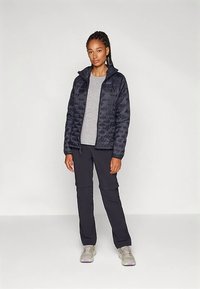 Casaco acolchoado azul-marinho com design matelassê, blusa cinzenta e calças cargo pretas. Sapatos claros com detalhes roxos completam o conjunto.