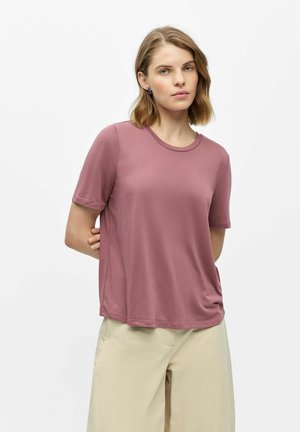 Vrouw met kort blond haar, gekleed in een mauve T-shirt met korte mouwen en beige broek, staand tegen een effen witte achtergrond.