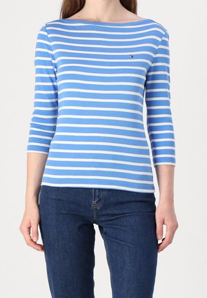 Kobieta ubrana w niebiesko-biały, poziomo paskowany, długi rękaw z małym logo na piersi, w połączeniu z ciemnoniebieskimi dżinsami.