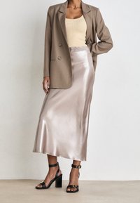 Beige blazer med dubbla knappar, ljus krämfärgad linne, satinlätt ljusrosa kjol och svarta klacksandaler med ankelremmar och guldaccenter.