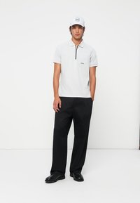 White polo shirt with a front pocket and black zipper detail, paired with loose black trousers and a white cap with a logo.