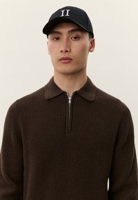 Jaunas vyras, dėvintis juodą kepuraitę su „II“ logotipu ir rudą ribuotą megztinį su užtrauktuku, stovintis priešais šviesų foną.