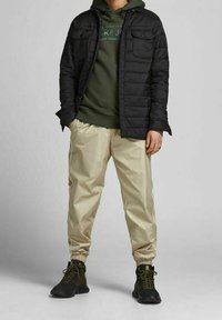 Chaqueta negra acolchada sobre una sudadera con capucha verde, pantalones beiges con cintura elástica y botas de senderismo negras con acentos verdes.