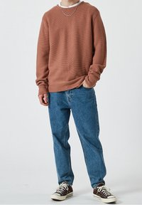 Person, der einen rostfarbenen Pullover, eine blaue Jeans, braune Sneakers mit weißen Schnürsenkeln und eine silberne Kettenhalskette trägt, steht mit einer Hand in der Tasche.