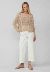 Beige gehäkelte Strickjacke über einem passenden Top, kombiniert mit weißen weit geschnittenen Hosen und Riemchensandalen. Schlichtes, strukturiertes Strickmuster.