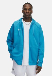Blaues Zip-Hoodie aus weichem Stoff, mit einer Kordelzugkapuze, Fronttaschen und einem gestickten Logo auf der Brust. Darunter ein schlichtes weißes T-Shirt.