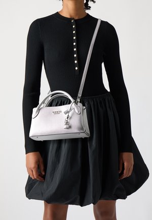 Personne portant une robe noire côtelée à manches longues avec des boutons, tenant un petit sac à main argenté avec une poignée supérieure et une bandoulière.