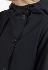 Chaqueta negra impermeable con un alto cuello, que presenta un cierre de botones y una textura suave. Incluye botones negros prominentes y un diseño minimalista.