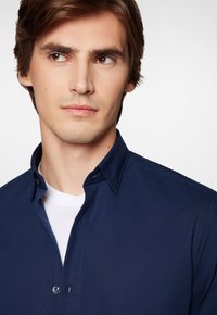 Granatowa koszula z guzikami, z usztywnionym kołnierzem, dwoma guzikami przy kołnierzu i kontrastowymi szwami. Noszona na białej koszulce.