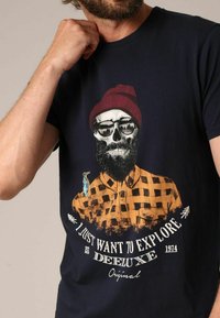 T-shirt bleu marine avec un graphisme d'un crâne avec une barbe, portant des lunettes de soleil et un bonnet rouge, sur un fond de chemise à carreaux orange.