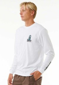 Camiseta blanca de manga larga con cuello redondo, que presenta un logo en teal y negro en el pecho y texto negro a lo largo de la manga izquierda.
