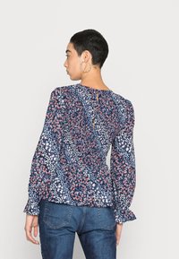 Blusa floral em azul escuro com padrões brancos e vermelhos. Apresenta mangas longas, corte peplum e uma abertura em forma de olho-chave na parte de trás.