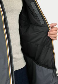 Intérieur d'une doudoune noire rembourrée avec une texture lisse, présentant des rayures multicolores le long de la fermeture éclair et un pull gris en dessous.