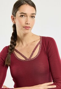 Burgundy long-sleeve top with a V-neck featuring textured decorative straps. Soft fabric with a smooth texture, minimal design.