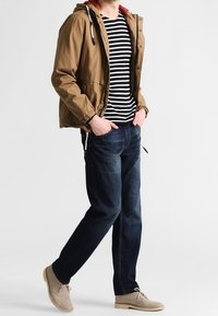 Homme portant une veste à capuche marron, une chemise rayée noir et blanc, un jean bleu foncé, des chaussures beiges, les mains dans les poches, marchant sur un fond blanc.