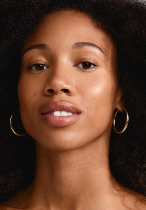 Gros plan d'une personne avec des cheveux bouclés et des boucles d'oreilles en or. La peau présente une texture lisse et un éclat naturel, avec des lèvres pulpeuses et des yeux marron.