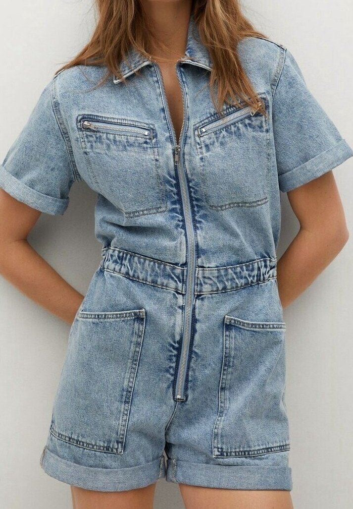 Femme portant une combinaison courte en jean bleu clair à manches courtes, fermeture éclair devant, manches retroussées et shorts, avec quatre grandes poches.
