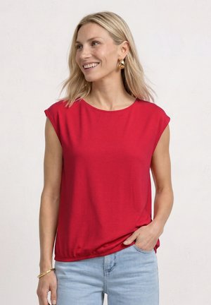 Femme souriante, portant un haut rouge vif sans manches avec un jean bleu clair, boucles d'oreilles créoles en or et un bracelet en or, main dans la poche.