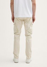 Beige Cargo-Hosen mit Seitentaschen, schmal zulaufenden Beinen und leichtem Stoff, kombiniert mit weißen Sneakern, von hinten betrachtet.