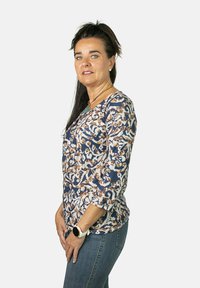 Camiseta de manga larga con un patrón floral abstracto en azul, beige y blanco. Presenta un escote en V y un corte holgado, combinada con jeans azules.