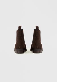 Bottes chevilles en daim marron avec des panneaux élastiques sur les côtés, des languettes au talon et une semelle en caoutchouc, vues de l'arrière sur fond blanc.