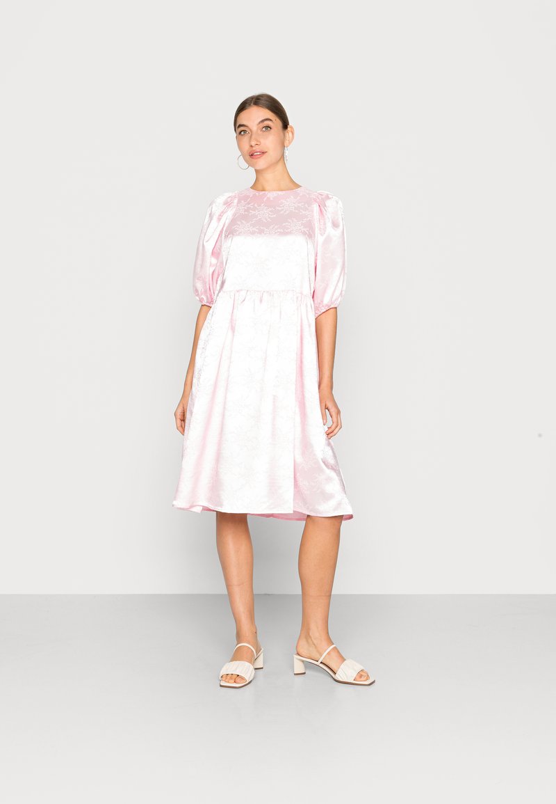 Vestido rosa de cetim com mangas bufantes e padrões florais, até ao joelho, com cintura franzida e uma textura suave, emparelhado com sandálias de salto bloco em bege.