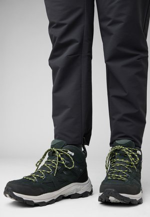 Grüne Wanderschuhe aus strukturiertem Stoff, mit schwarzen Akzenten und leuchtend gelben Schnürsenkeln. Getragen mit dunklen, flexiblen Hosen, die eine schmale Passform haben.