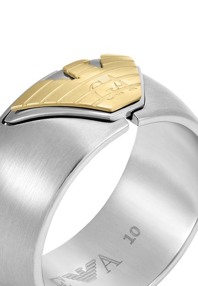 Bague en métal avec une finition argent brossé et un accent doré présentant un design ailé. Marquages gravés sur l'intérieur de la bande.