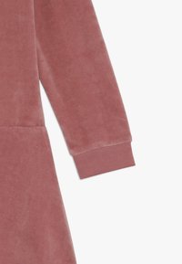 Růžové sametové šaty s dlouhými rukávy, žebrovanými manžetami a hladkým povrchem; mají klasický siluetu a jemný lesk.