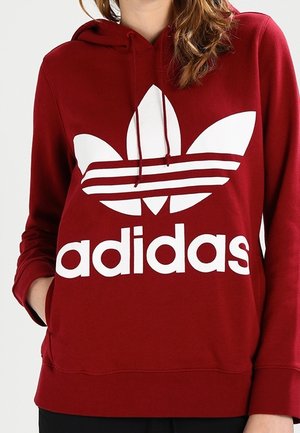 Osoba nosząca czerwoną bluzę z kapturem z dużym białym logo Adidas i motywem trójlistnej koniczyny z przodu, ręce w kieszeniach.