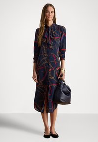 Γυναίκα που φοράει φόρεμα σε ναυτικό μπλε με σχέδιο αλυσίδας και ζωνάκι, κρατώντας σκούρα δερμάτινη τσάντα, μαύρες μπαλαρίνες και χρυσά βραχιόλια, στέκεται σε απλό φόντο.