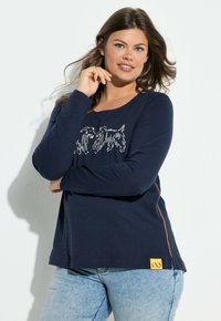 Maglietta a maniche lunghe blu navy con un'illustrazione bianca di un cane sulla parte anteriore, con un accento arancione sul lato e un'etichetta con il logo giallo all'orlo.