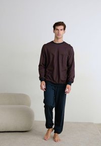 Haut à manches longues à motifs marron avec des ornements noirs, associé à un pantalon bleu marine. Le mannequin se tient pieds nus sur un tapis doux aux tons neutres.