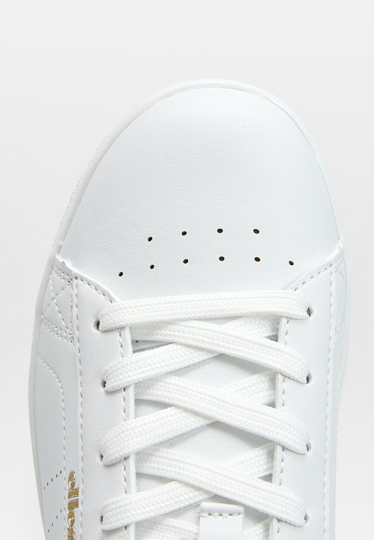 Weißer Ledersneaker mit flachen Schnürsenkeln, Perforationen an der Zehe und einem goldenen Akzent in der Nähe der Sohle. Hat eine glatte, strukturierte Oberfläche.