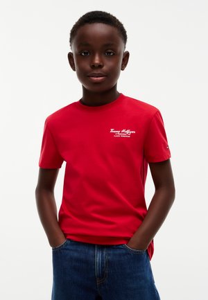 Garçon debout les mains dans les poches, portant un t-shirt rouge Tommy Hilfiger et un jean bleu sur un fond blanc simple.