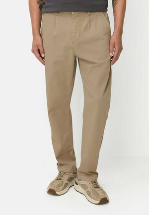 Mann trägt beige gerade geschnittene Hosen und passende beige Sneakers, steht vor einem einfarbigen Hintergrund.