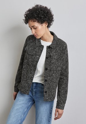Chaqueta de tweed blanco y negro con cuello tipo solapa, bolsillos delanteros y cierre de botones, combinada con una blusa blanca de canalé y jeans azules.