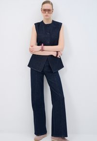 Top sin mangas de mezclilla oscura con botones en el frente y pantalones acampanados a juego. Los accesorios incluyen una pulsera rosa y gafas de sol grandes. Fondo liso.