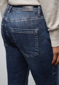 Jeans en denim bleu moyen avec une texture lisse, dotés de poches arrière et d'une étiquette noire sur la ceinture. Design ajusté.