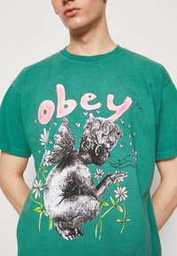 Zelené bavlněné tričko s grafickým designem cheruba s křídly, květinami a slovem "poslouchej" v růžové barvě nahoře.