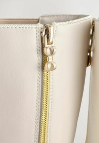Beige neplederen tas met een gouden rits en een decoratieve trekker. De rits is centraal geplaatst langs de rand en voorzien van stiksels.
