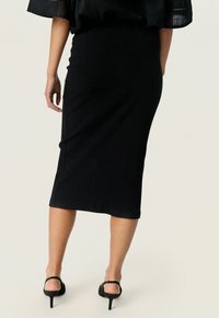 Jupe midi noire à côtes avec une silhouette ajustée, présentant une texture lisse et un léger éclat, associée à des sandales noires à talons hauts.