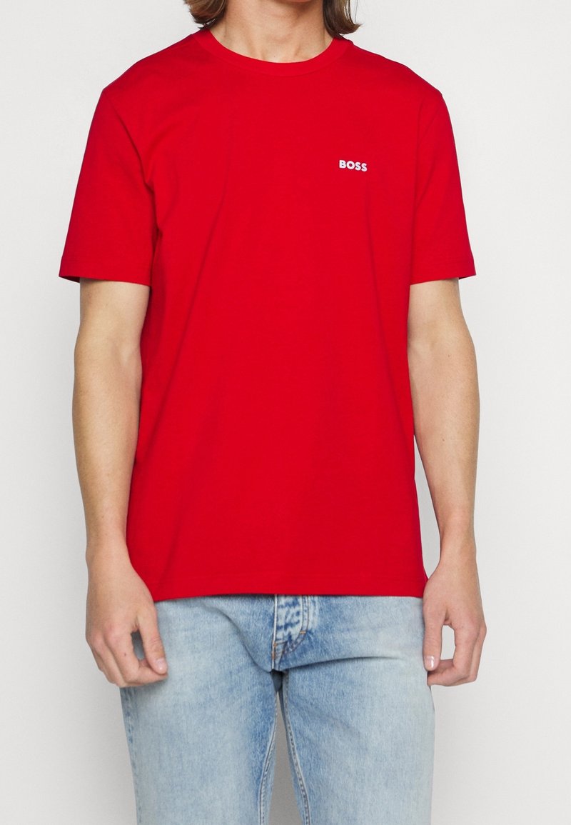 Personne portant un t-shirt rouge uni avec le logo "BOSS" sur la poitrine et un jean bleu clair, debout devant un fond blanc.