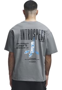 Personne aux cheveux courts portant un T-shirt gris avec l'inscription « INTROSPECT Yourself » et un graphisme d'un vieux téléphone et de petits rats bleus dans le dos.