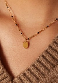 Collier en chaîne dorée avec des perles noires, présentant un pendentif ovale. La chaîne a une finition texturée et repose sur une peau bronzée.