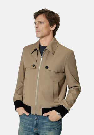 Mężczyzna ubrany w beżową kurtkę na suwak z czarnymi mankietami i klapami kieszeni, w niebieskich dżinsach, stojący z rękami w kieszeniach.