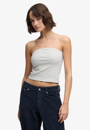 Kvinde med kort brunt hår iført en stropfri grå og hvid stribet cropped top og mørkeblå ribvelourbukser mod en hvid baggrund.