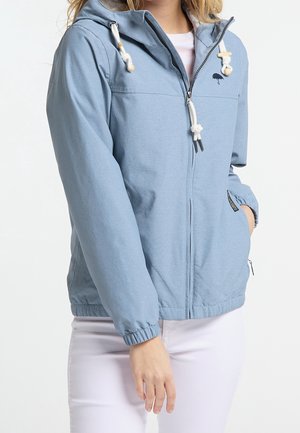 Vrouw draagt een lichtblauwe hoodie met rits, witte koordjes, elastische manchetten en hand in zijzak, gecombineerd met een witte broek.