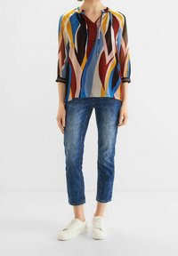 Blusa a maniche lunghe multicolore con scollatura a V, in tessuto trasparente e motivo astratto; abbinata a jeans blu a vita alta e sneakers bianche.