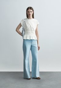 Mujer con una blusa blanca ajustada en la cintura, vaqueros de pierna ancha azul claro con adornos, zapatos de estampado de leopardo y un bolso de hombro plateado.